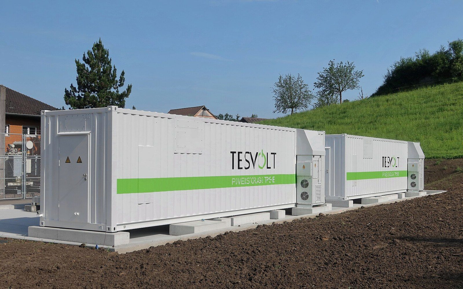 Two white battery storage containers on concrete pads beside a fenced compound